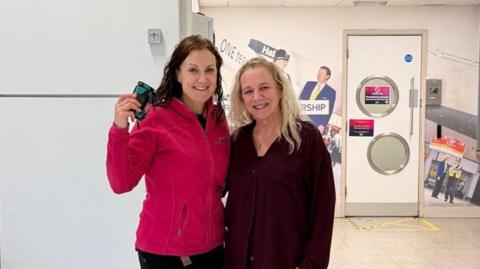 A woman with dark hair and a pink top holding up a small dark object beside a woman with blonde hair and a dark top, both standing in a large room with white walls and a tiled floor