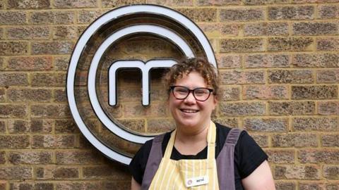 Beth Hodgson, who has curly brown hair, stands in front of the brick wall with a white MasterChef logo fixed to it. She is wearing a yellow and white pinstriped apron with her name shown on a tag. Beneath the apron she is also wearing a black T-shirt and navy overalls.