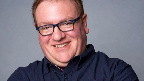 Adrian Hobart has short blond hair and black glasses. He is also wearing a dark blue shirt