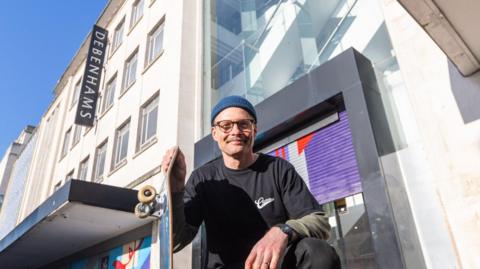 Tim standing outside the old entrance to the Debenham's department store crouching down with his skateboard smiling at the camera.