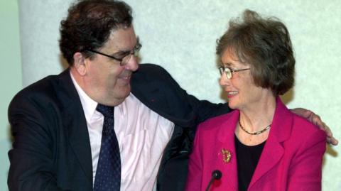 John Hume has his arm around Bríd Rodgers as they smile at each other. Hume has black curly hair and is wearing glasses, a dark suit, pink shirt and navy tie. Rodgers is also wearing glasses and is wearing a pink jacket and black top. She has a gold brooch pinned to her jacket.