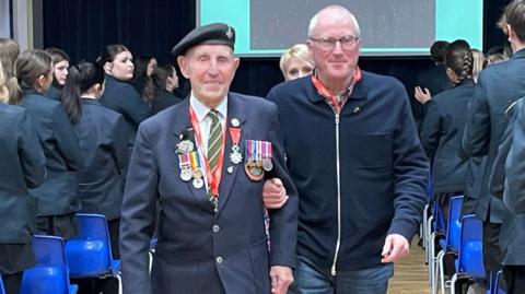 Two men walking down the aisle of a school hallway. One is wearing a dark blazer with multiple military medals and a beret. The other man is dressed in a zip-up jacket and jeans, walking alongside and supporting the other man by holding his arm. In the background, a large projection screen displays a message and an image. Either side of the men are male and female students dressed in  dark coloured school uniform. 