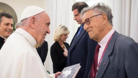 Pope Francis, dressed all in white, is holding the book and smiling as he talks to David Willey, who is also smiling.