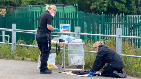 Two female police officers wearing surgical masks examine items next to a camping table holding plastic evidence bags