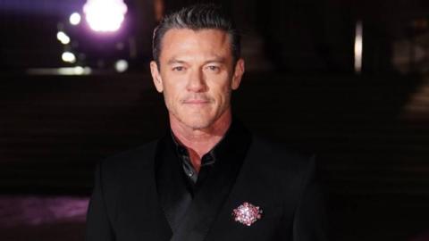 A man with greying hair and a moustache, wearing a black velvet suit with a pink brooch on his lapel.