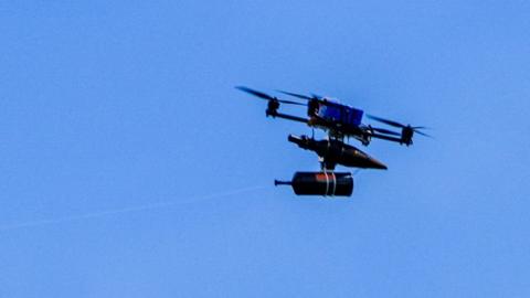 A fibre-optic drone flies in a clear blue sky across the border from Lebanon into Israel. It has a spool of fibre optic wire handing under a chassis, supported by four propeller blades around a blue rectangular battery.