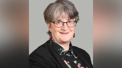 A woman with short black and grey hair looks into the camera, wearing black-rimmed glasses. She has a dark jacket on over a black shirt with a light floral pattern.