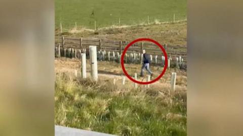 A screengrab of a mobile phone video showing a man in a field next to a road.