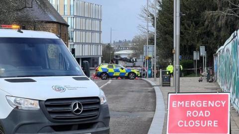 A van and a red emergency road closure sign on a road. In the background are a police car and a person in a high-vis jacket.