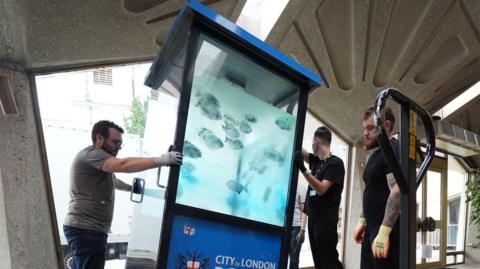 Three workers are moving a City of London Police box which has fish painted on its windows. 
