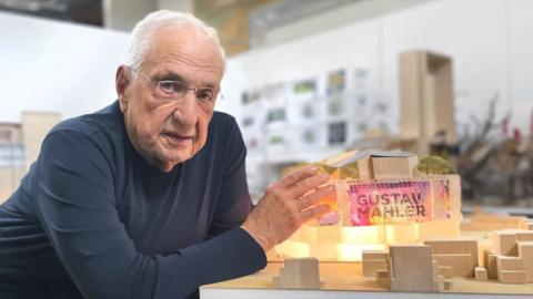 A person wearing a dark long-sleeve shirt is leaning on a table, presenting a detailed architectural model, featuring illuminated sections, miniature trees, and geometric structures in a modern studio setting.