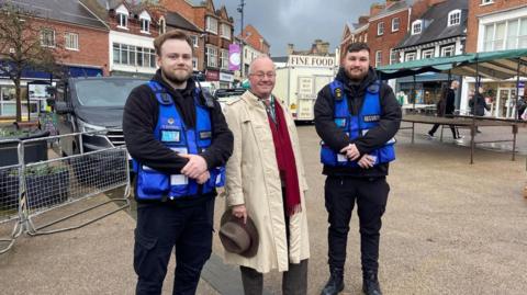 PCC Rupert Matthews standing between two blue uniformed security guards