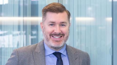 A photo of Alan Pease. He is pictured facing the camera and smiling. Behind him are windows that have been covered with blinds. He is wearing a grey suit with a light blue shirt and a navy tie. He has light brown, short hair and has grey stubble. 