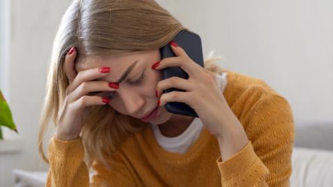 Woman wearing a yellow sweater has her hands on her head while holding up a phone to her ear. She looks stressed.