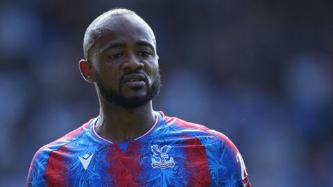 A head and shoulders shot of Jordan Ayew, wearing Crystal Palace's home strip, with the player looking downwards with his mouth slightly open