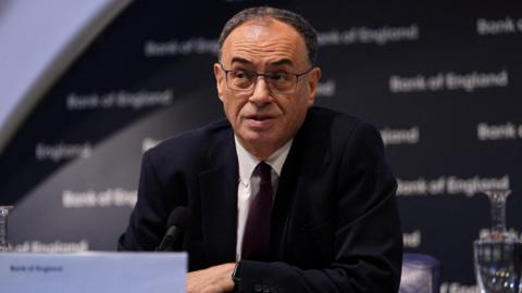 Andrew is wearing a dark suit jacket over a white shirt and black tie. He is sitting on a conference table with a dark background reading: Bank Of England.