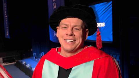 Lee Radford smiling at the camera. He is wearing a graduate's cap and gown. His cap is black with red tassels and his gown is red and blue. He is standing in a large theatre on a platform
