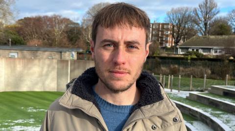 A man with brown hair and stubble looking at the camera. He is wearing a beige coat and standing in an open air theatre which has snow on the steps.