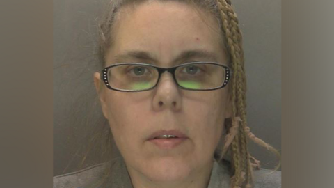 A police mugshot of a woman with her blonde hair braided on one side, wearing dark-framed glasses and a grey top
