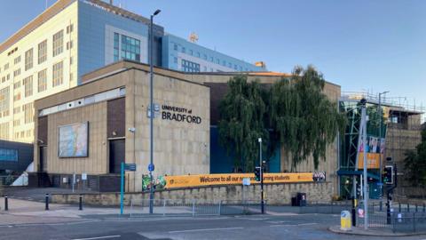 A sandstone building with black lettering spelling the University of Bradford with a pedestrian crossing in front. 