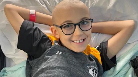 Ezra is laying on a hospital bed and is looking directly at the camera and is smiling. His arms are behind his head and he is wearing a black, grey and yellow football shirt.