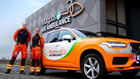 Two people are stood next to one of the rapid response cars, in front of the Magpas building. They wear bright orange cargo pants with and orange and black fleece. It matches the orange, green and which car.