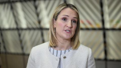 Helen McEntee is talking. She has shoulder-length blonde hair and is wearing a cream coloured jacket over a stripy top. Behind her out of focus a glass wall can be seen.