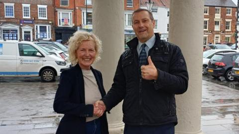 Denise Howard and Jon Dimberline shake hands while smiling at the camera. Denise Howard has short blonde curly hair and is wearing a navy blazer, a neutral-coloured t-shirt and blue jeans. Jon Dimberline is wearing a black puffer coat, a light blue patterned shirt and a blue checked tie. He is giving a thumbs- up with his other hand. There is a car park behind them full of vehicles.