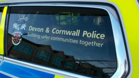 The window of a police car with the words 'Devon & Cornwall Police Building safer communities together'. There is a badge on the left. 