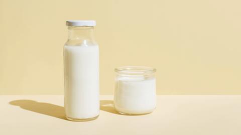 A glass bottle of milk and small glass of milk next to it. With a pale yellow background. 