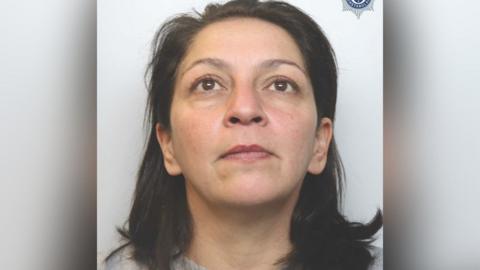 A police mugshot of a woman with long black hair and a grey top. She is looking up. 