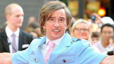 Steve Coogan attends the Alan Partridge: Alpha Papa London Premiere dressed as Alan Partridge. He wears a light blue suit with a white shirt underneath and a pink tie. He has long hair and onlookers can be seen behind him.