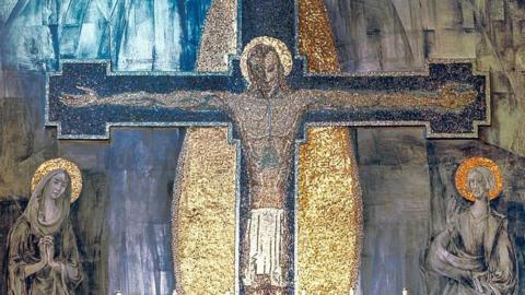 Jesus is depicted with a golden halo with his arms stretched and pinned to the cross with Mary and St John - also with golden halos - praying on both sides. The cross is surrounded by a dazzling golden oval.