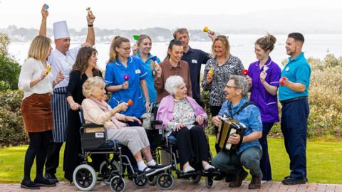 Two older women are sat in wheelchair in front of a lush garden. They are both looking at a man who is crouched down in front of them and playing the accordion. Behind them are 10 others, most of whom appear to be staff from the care home. Many of the people are holding small tambourines or bells. To the left, a chef, wearing a traditional chef's hat, raises both hands in the air. 