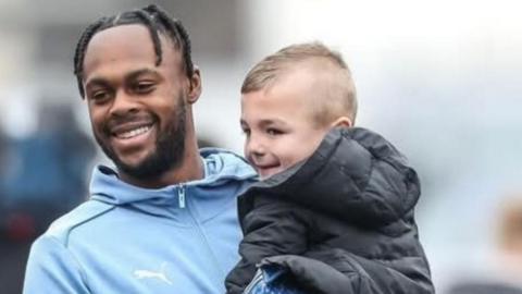 Striker Ricky-Jade Jones in a pale blue zip-up training top, carries Frankie, who is wearing a black coat. Both are smiling.