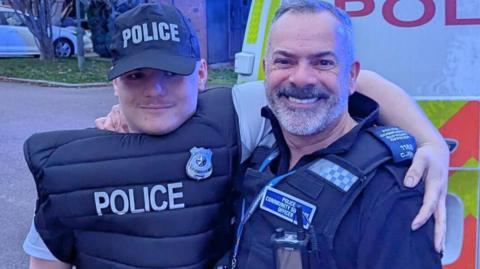 Ryan Sissons wearing a police cap and vest with PCSO Chris Jones in his uniform standing in front of a police van