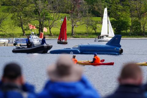 The restored Bluebird K7, which crashed killing Donald Campbell in 1967, takes to the water in Coniston, Cumbria, during the Bluebird K7 Festival. Spectators are in the foreground, out of focus, and Bluebird with support craft are on the water. Beyond Bluebird are two sailing boats, one with a white sail and one with red. Bluebird is a long, low, blue vessel, similar in design to an old fashioned racing car. Beyond the water are green fields and scattered trees.