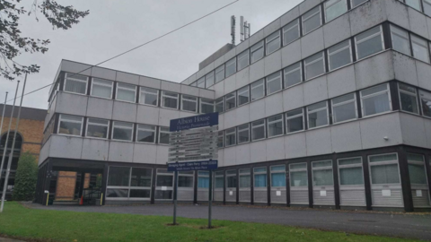 A white four-storey building is L-shaped and has a sign outside the building which says Albion House. There is a door at the left side of the building.