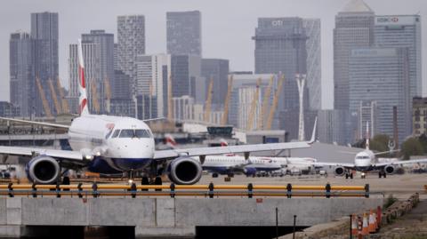 A British Airways plane sitting at the airport. The tall buildings of the Canary Wharf financial district can be seen in the background.
