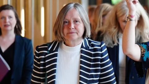 Helen McDade, who has short grey hair, looks at the camera as she walks along a corridor. She is wearing a stripy top over a white jumper 