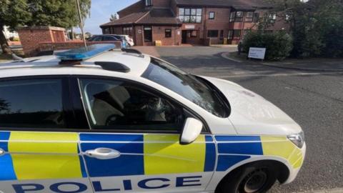 A white, yellow and blue police care dominates the front of the picture. In the background a brown brick building can be seem. There is a white sign but it cannot be read. 