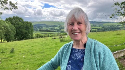 Dina Smith in turquoise cardigan leaning on a fence in green valleys