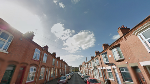 A street view of Raymond Road in Leicester. It depicts terraced properties on both sides of the road and parked cars.