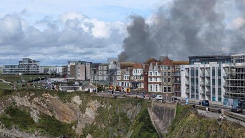 A large plume of black smoke comes from the roof on an multi-storey Victorian hotel on a Cornish clifftop, surrounded by others.
