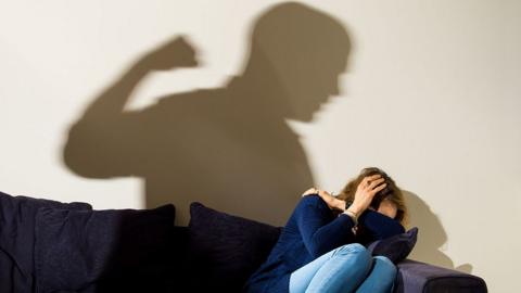 Generic images showing a woman in jeans and blue jumper cowering on a sofa with her hands covering her head while the shadow of a man with his fist raised is on the wall behind her