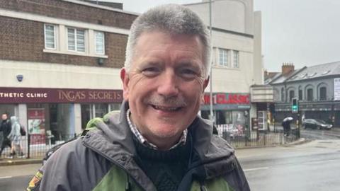 Peter Smalley standing in a street smiling at the camera on a rainy day. 
