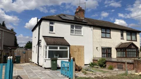 A white semi-detached house with a dark roof and a blue gate leading to a paved drive. Part of the front of the house is covered with wood.