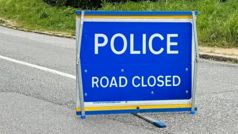 A blue sign on a road with the words 'POLICE' and 'ROAD CLOSED' below it. It has a thin yellow trim at the top and bottom. The road is grey and there is grass in the background. 