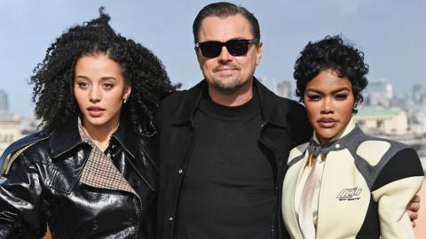 Chase Infiniti, Leonardo DiCaprio and Teyana Taylor attend the London photocall for One Battle After Another in London in September. Infiniti is wearing a black shiny jacket, DiCaprio has a black jacket and T-shirt on and Taylor has on a racing-style cream, grey and black jacket.