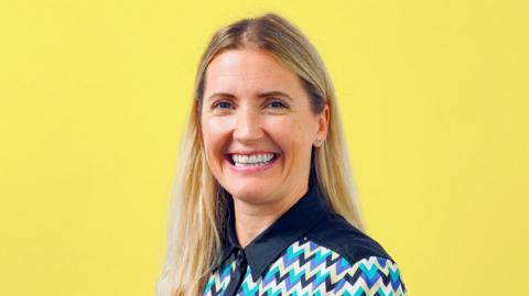 Professor Donna Whitehead looks directly at the camera and smiles. The background of the image is a solid yellow colour and she is wearing a top with a turquoise, purple, yellow and black zig zig pattern on and she has long blond hair.